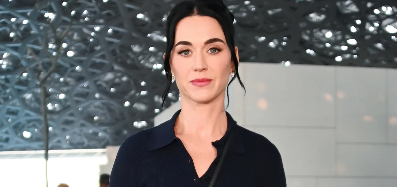 Katy Perry Katy Perry z ciemnymi włosami spiętymi w kok, w granatowej koszuli, na tle nowoczesnej ściany z geometrycznym wzorem.
