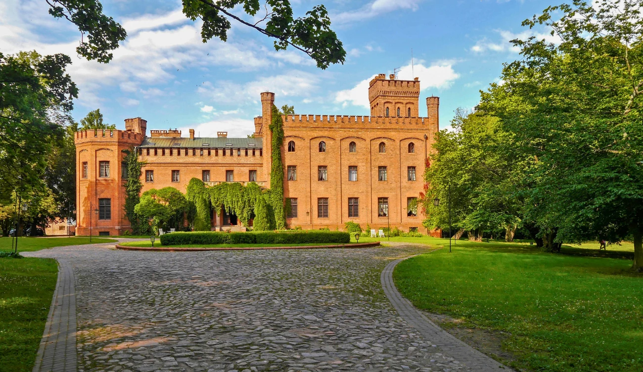 Zamek Sobieskiego w Rzucewie Ceglany zamek z charakterystycznymi wieżyczkami i blankami otoczony zadbanym parkiem z bujną zielenią, brukowaną alejką prowadzącą do głównego wejścia oraz wysokimi drzewami po obu stronach.