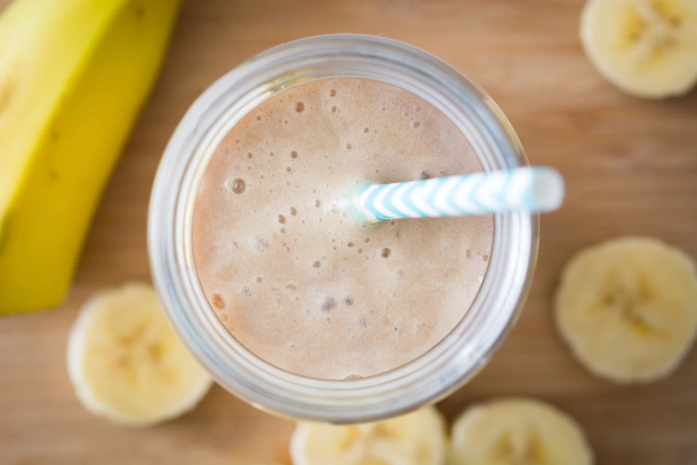 Bananowa kawa mrożona to hit na cieplejsze dni. Bez trudu zrobisz ją we własnej kuchni Szklanka z bananową kawą mrożoną z niebiesko-białą słomką, plastry banana i cały banan na desce.