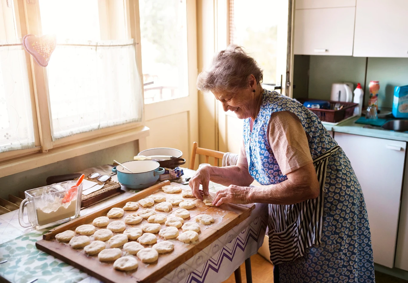 Bądź bardziej jak "nonna". Trend okrzyknięto "najbardziej sensownym w historii" Starsza kobieta w fartuchu formuje okrągłe ciasteczka z ciasta na desce w jasnej kuchni przy oknie.