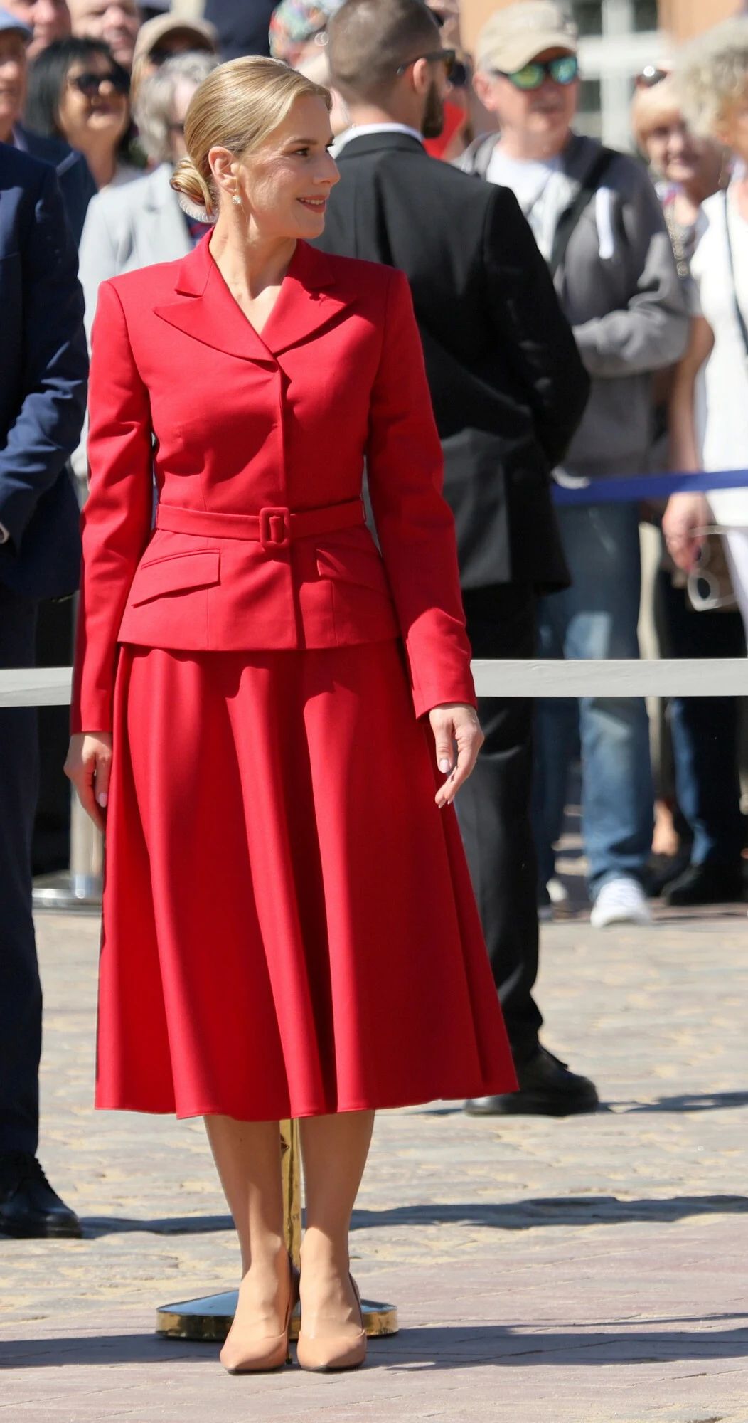 Kobieta w eleganckim czerwonym garniturze z rozkloszowaną spódnicą i beżowych butach na tle grupy ludzi podczas wydarzenia na świeżym powietrzu.