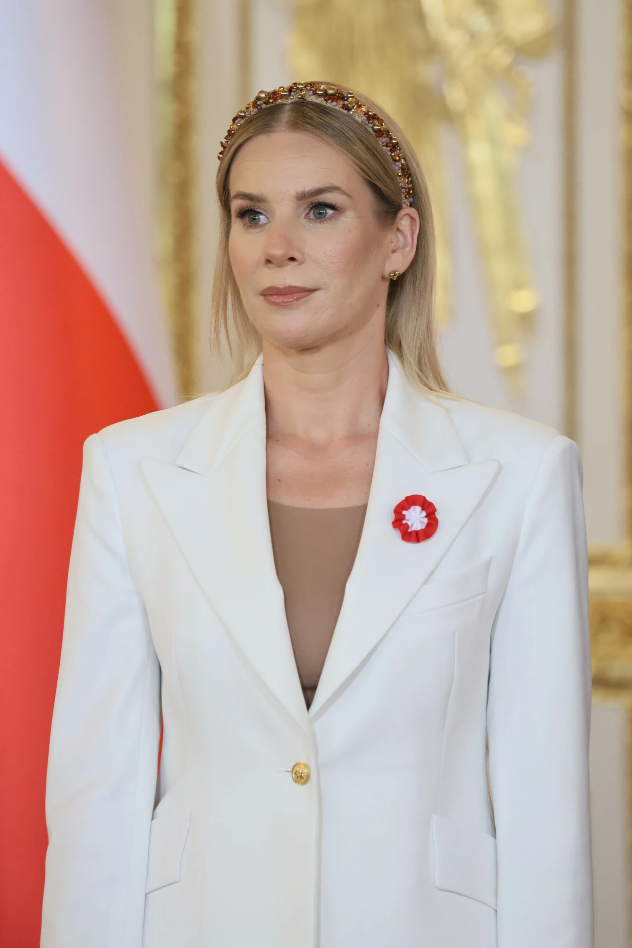 Kobieta w eleganckiej białej marynarce z przypiętą biało-czerwoną rozetką, ozdobna opaska na jasnych włosach, delikatny makijaż, złote detale w tle.