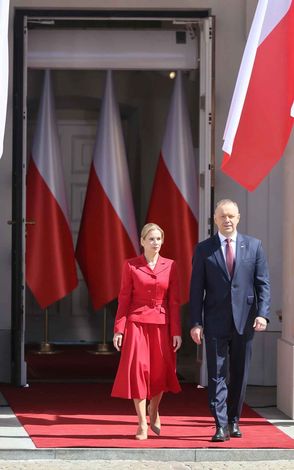 Dwoje elegancko ubranych osób idących po czerwonym dywanie przed budynkiem, w tle widoczne polskie flagi.
