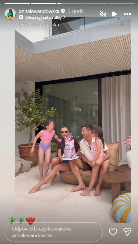 Anna Lewandowska i Robert Lewandowski z córkami, Klarą i Laurą Czteroosobowa rodzina siedzi razem na tarasie na beżowej kanapie, trzymając kolorowy tort urodzinowy ozdobiony motywem kwiatów i liczbą 7.