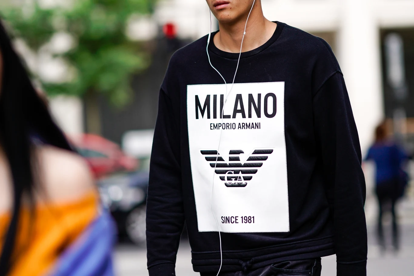 Mężczyzna w czarnej bluzie Emporio Armani z napisem 'MILANO' oraz dużym logo marki, ze słuchawkami w uszach, w miejskim otoczeniu.