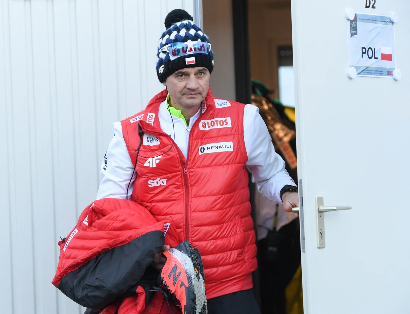 Mężczyzna w czerwonej kamizelce sportowej i czapce wychodzi z białego pomieszczenia, trzymając kurtkę oraz narty, na drzwiach widoczna kartka z napisem POL i flagą Polski.