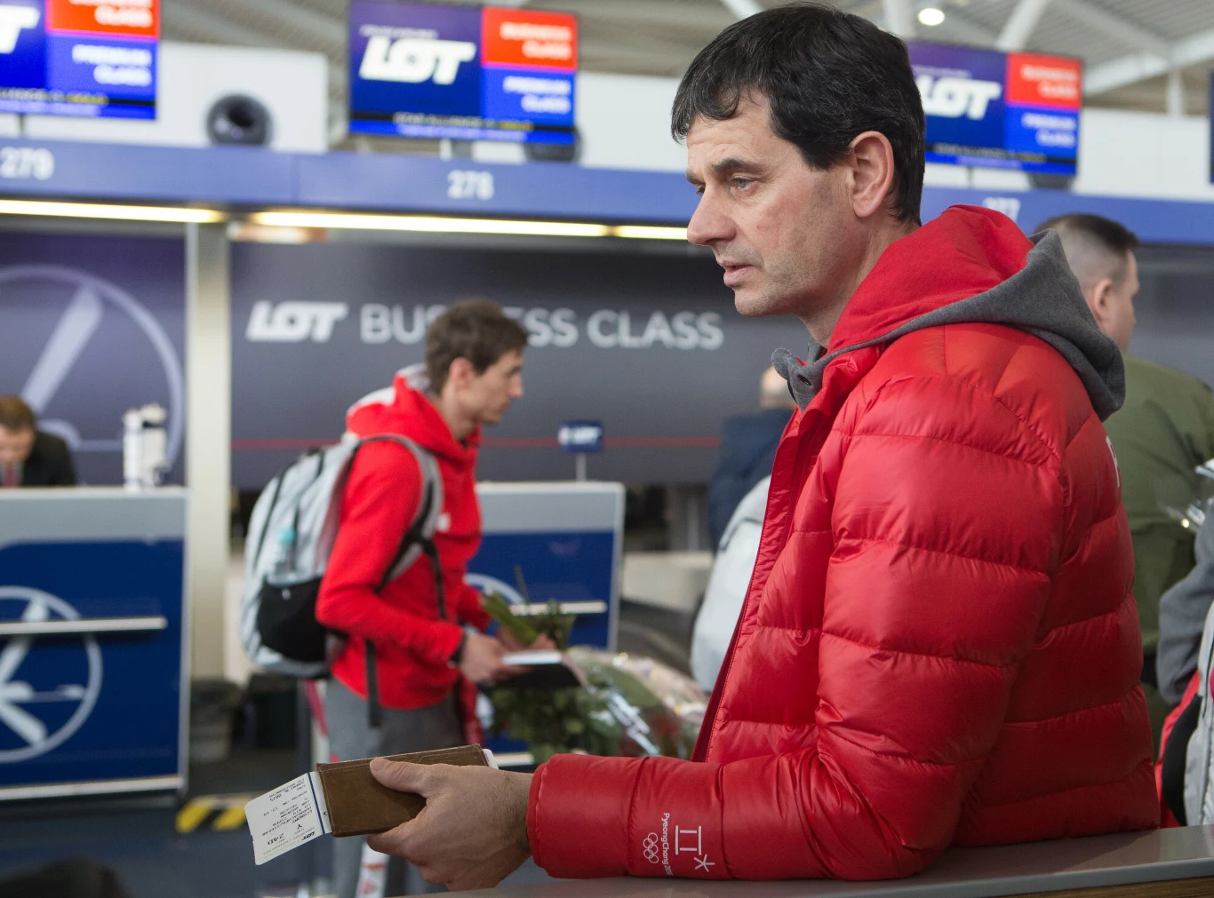 Stefan Horngacher Mężczyzna w czerwonej kurtce stoi przy stanowisku odprawy biletowej LOT Business Class, trzymając dokumenty podróżne w dłoni, w tle widoczne są inne osoby oraz ekrany informacyjne linii lotniczych.