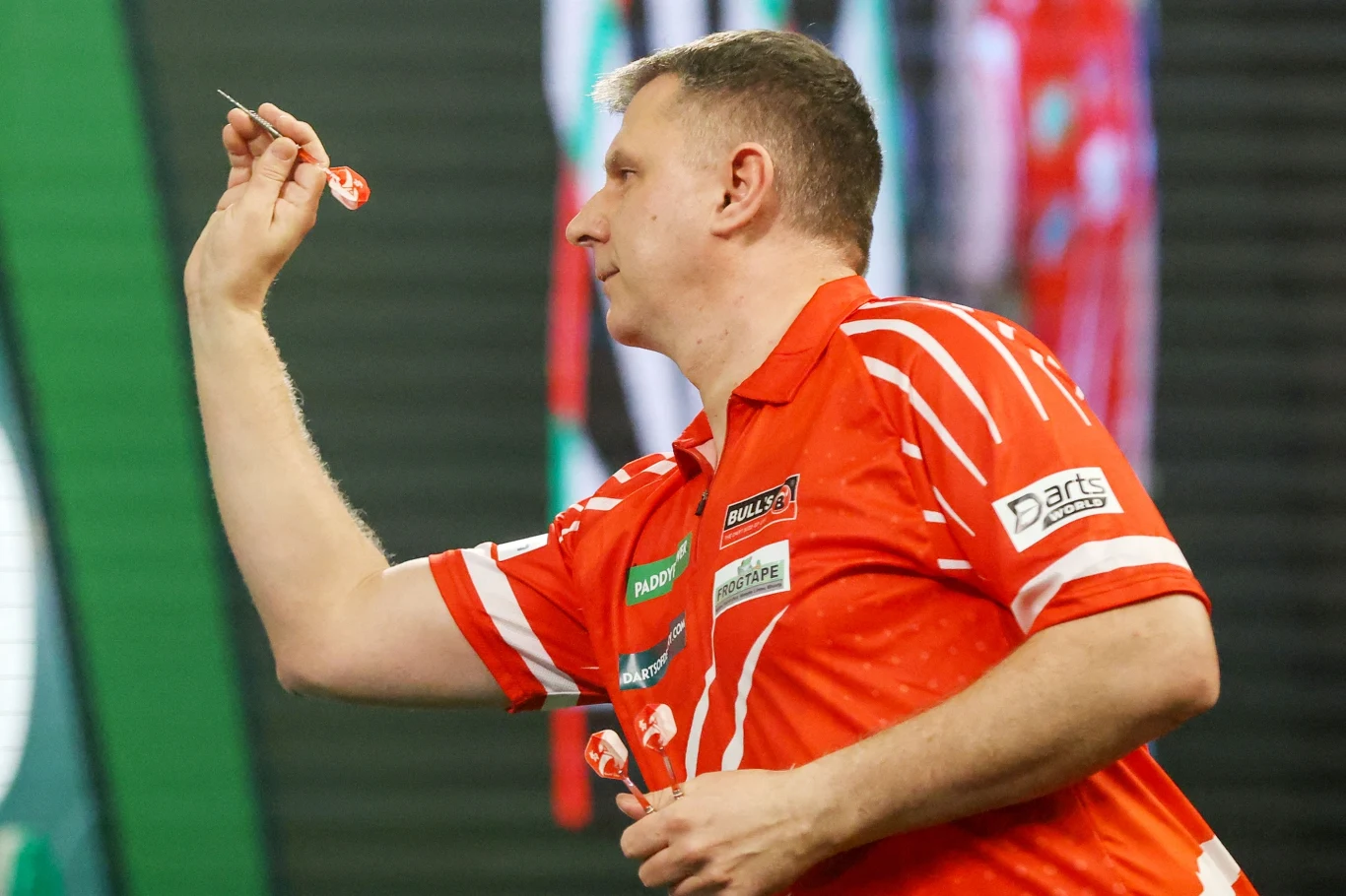 Mężczyzna w czerwonej koszulce sportowej koncentruje się, trzymając rzutkę w jednej ręce i przygotowując się do oddania rzutu podczas zawodów w darta.