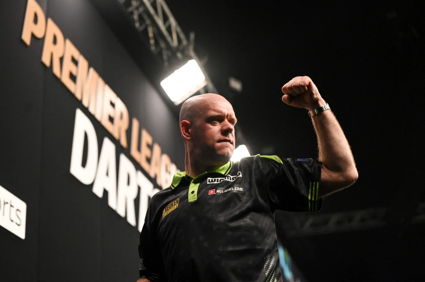 Zawodnik darta w ciemnej koszulce z uniesioną w geście triumfu ręką na tle napisu PREMIER LEAGUE DARTS.