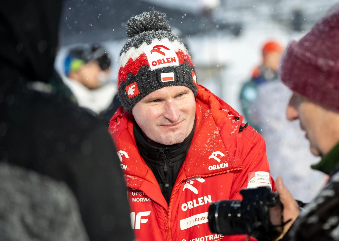 Maciej Maciusiak Maciej Maciusiak w czerwonej kurtce i czapce ORLEN rozmawia z osobą na śniegu podczas wydarzenia sportowego.