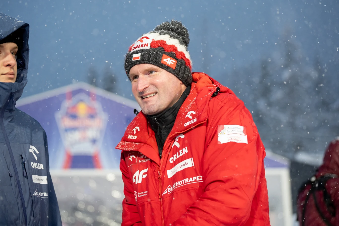 Maciej Maciusiak Mężczyzna w czerwonej kurtce i czapce z biało-czerwonym akcentem, stojący na śniegu podczas zimowej imprezy sportowej, w tle zamazany logotyp oraz inne osoby ubrani w ciepłą odzież.