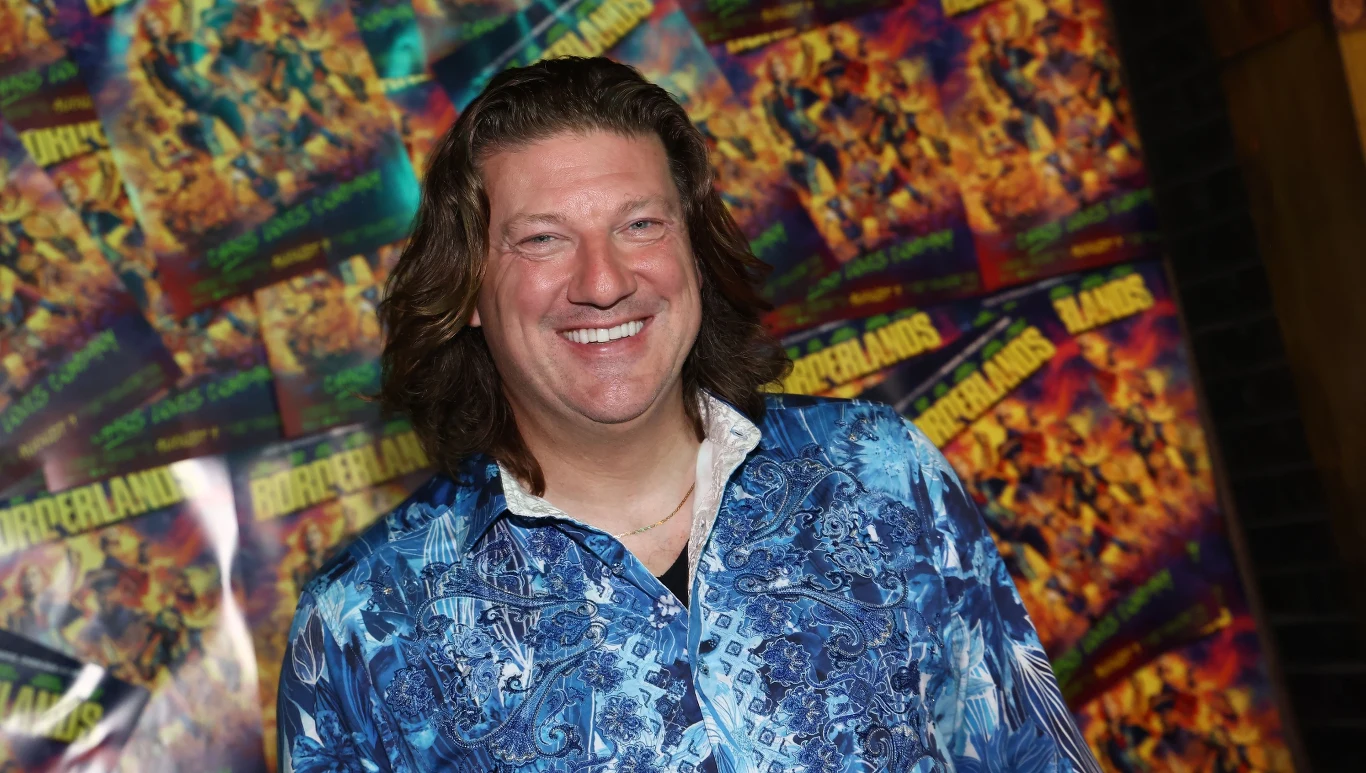 Randy Pitchford Randy Pitchford w niebieskiej koszuli z wzorem, stoi przed plakatami gry Borderlands.