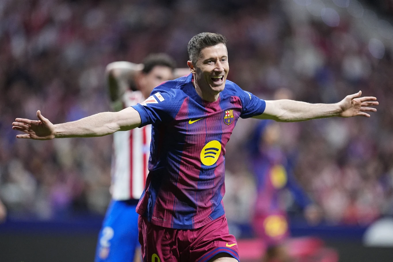 Piłkarz w bordowo-granatowej koszulce FC Barcelony z szeroko rozłożonymi ramionami świętuje zdobycie gola na boisku podczas meczu piłkarskiego.