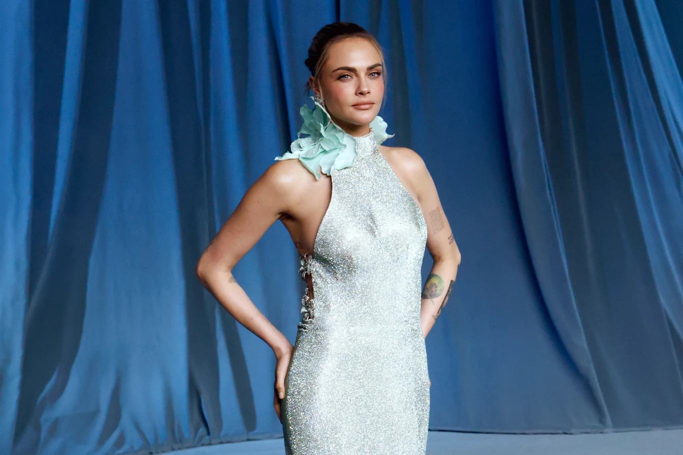 Cara Delevingne rozpoczyna muzyczną karierę Cara Delevingne w błyszczącej, dopasowanej sukni z odsłoniętymi ramionami, przy szyi jasna dekoracja.