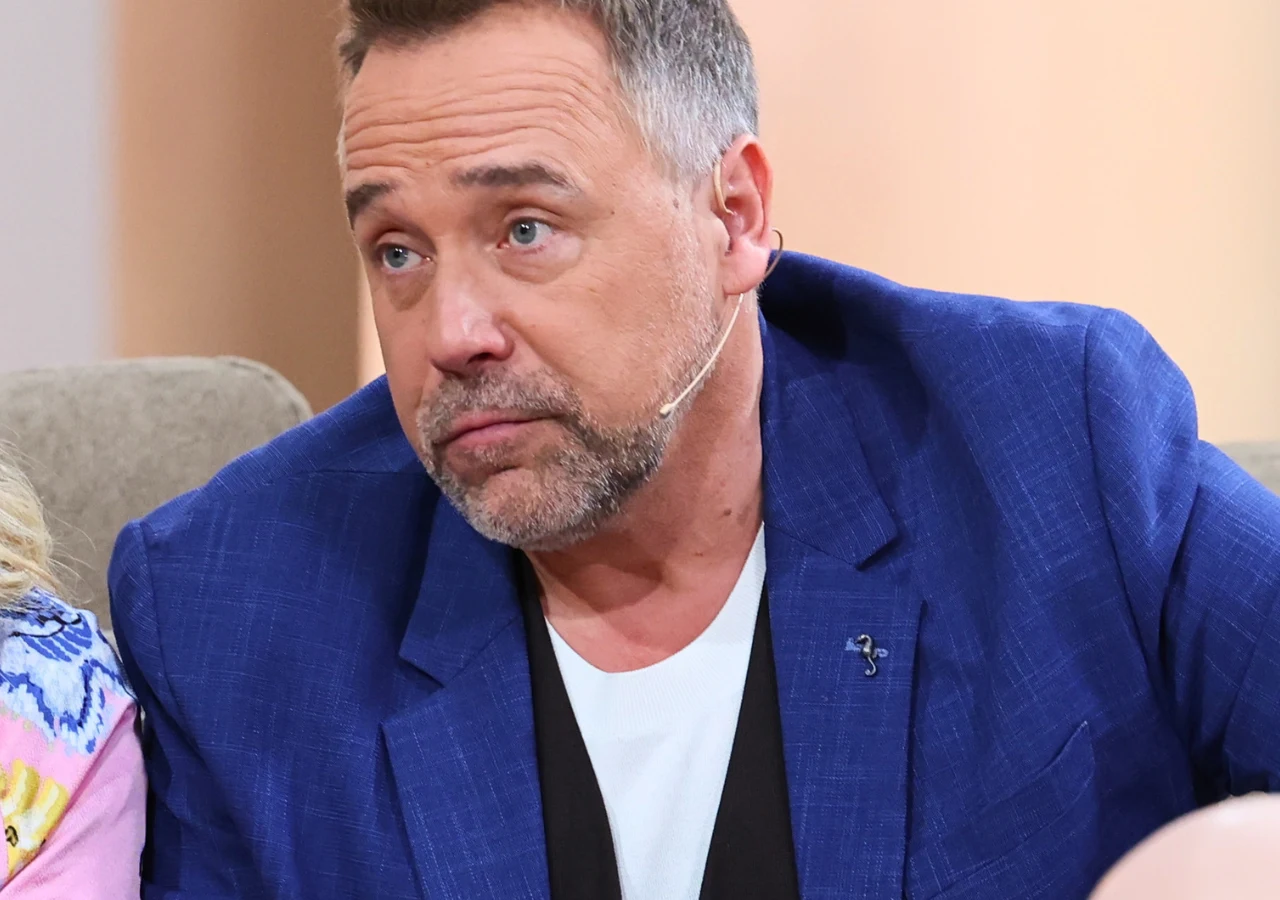 Łukasz Nowicki Prezenter Łukasz Nowicki w niebieskiej marynarce z przypiętym mikrofonem siedzi na kanapie i patrzy w bok.