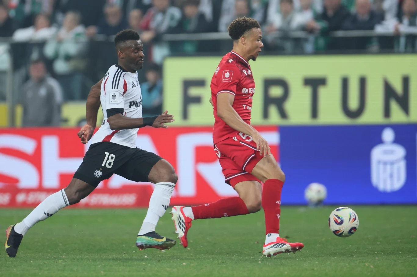 Dwóch piłkarzy w dynamicznym biegu za piłką podczas meczu, zawodnik w białej koszulce ściga zawodnika w czerwonym stroju na tle stadionu pełnego kibiców.
