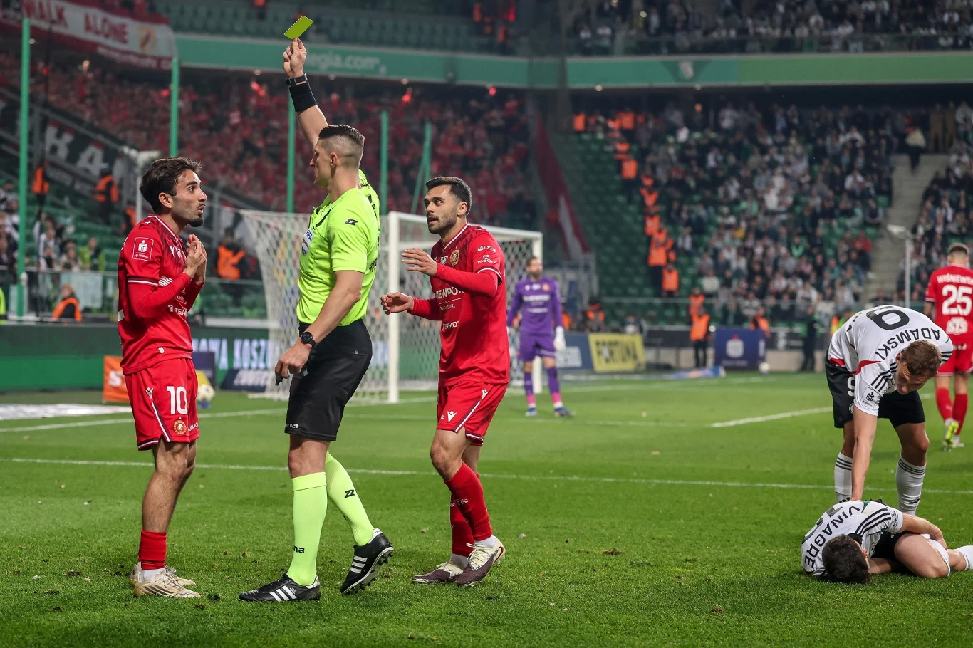 sędzia pokazuje zawodnikowi w czerwonym stroju żółtą kartkę, obok drugi piłkarz w czerwieni protestuje, a na murawie leży zawodnik drużyny przeciwnej trzymający się za nogę