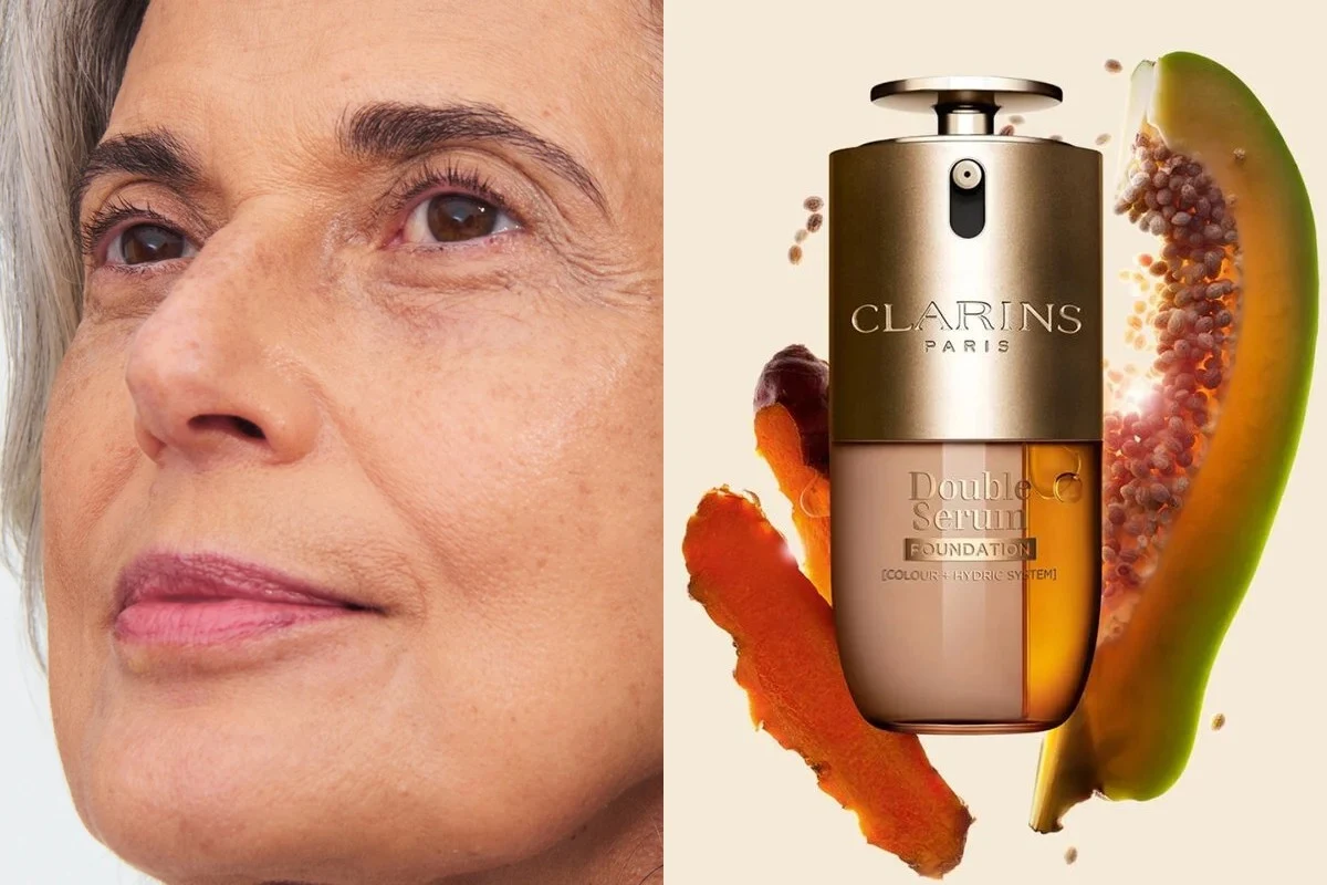 Zbliżenie twarzy starszej kobiety z naturalnym makijażem obok eleganckiego flakonu podkładu Clarins oraz przekrojonej papai i plam składników aktywnych.