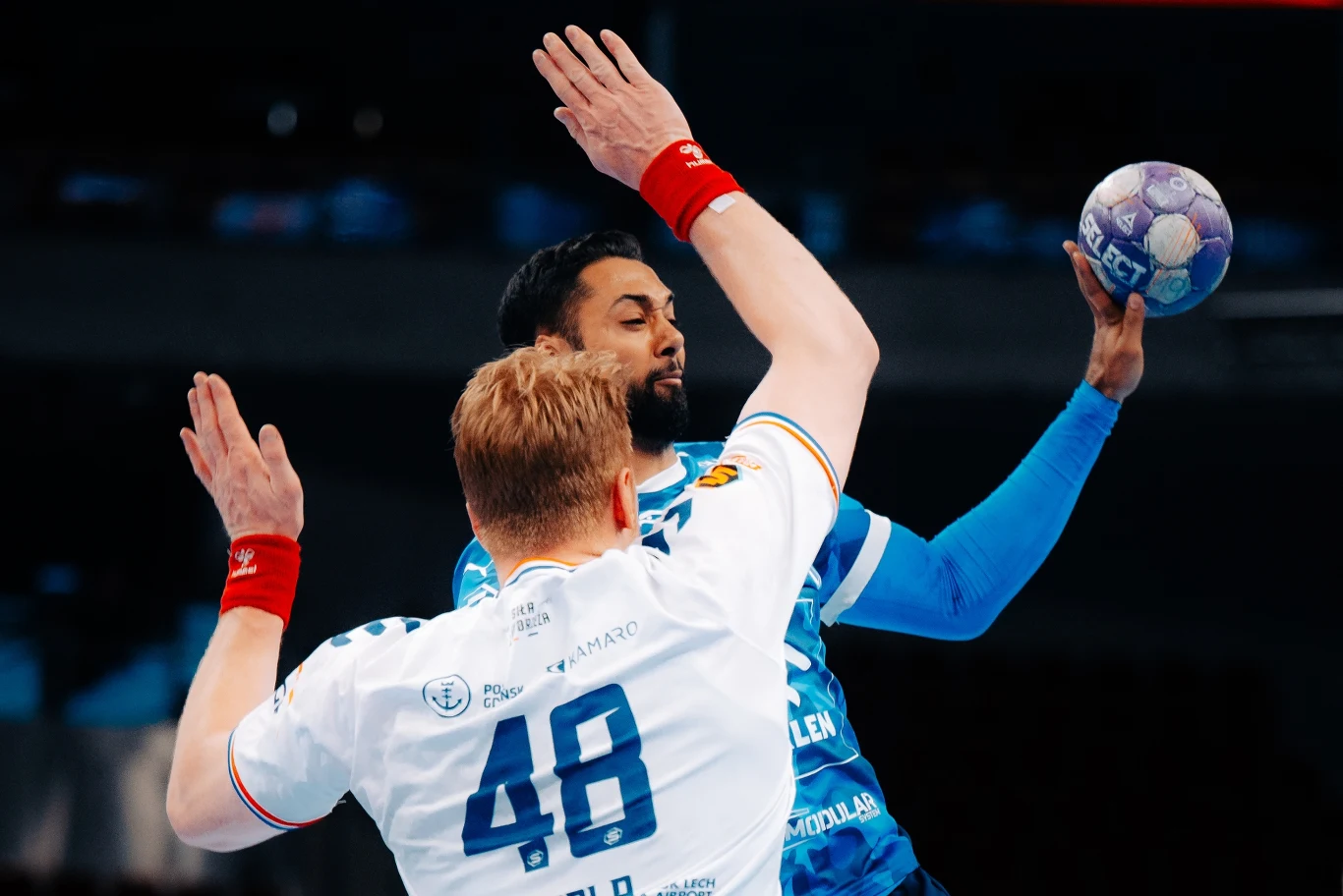 PGE Wybrzeże Gdańsk – ORLEN Wisła Płock Dwóch zawodników piłki ręcznej w dynamicznej walce o piłkę, gracz w niebieskim stroju unosi rękę z piłką, próbując rzutu, podczas gdy zawodnik w białym stroju stara się go zablokować.