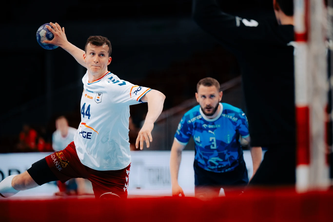 PGE Wybrzeże Gdańsk – ORLEN Wisła Płock Zawodnik drużyny piłki ręcznej przygotowujący się do rzutu na bramkę, skoncentrowany bramkarz w niebieskim stroju obserwuje tor lotu piłki.
