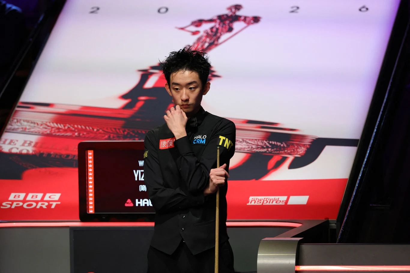 Zawodnik snookera w czarnym stroju z bilą do gry w ręku stoi zamyślony obok stołu, na tle dużej grafiki stołu snookerowego i logo BBC Sport.
