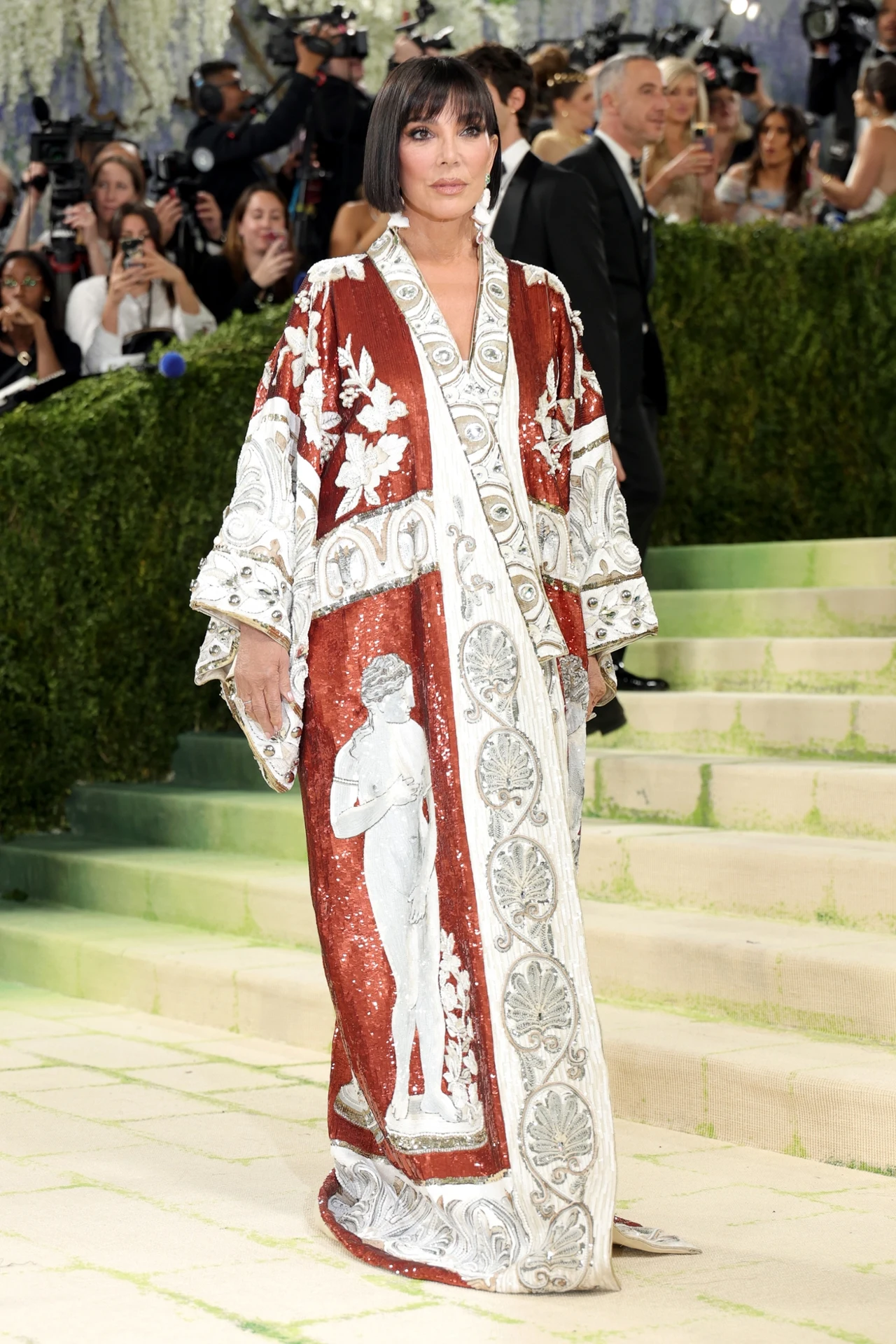 Stylizacja, fryzura i twarz - wszystko w look'u Kris Jenner było magnetyczne Kobieta w efektownym, długim kimono z bordowo-białym wzorem oraz motywami postaci i roślin, stojąca na schodach podczas oficjalnego wydarzenia wśród licznych fotografów.