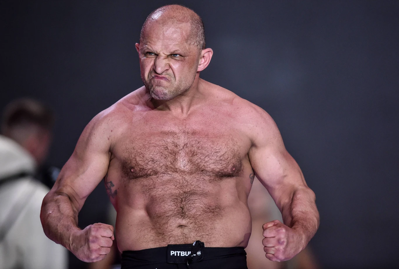Jacek Murański Jacek Murański, umięśniony łysy mężczyzna w spodenkach sportowych, napina ramiona na ciemnym tle.
