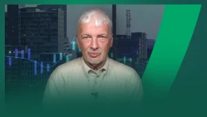 Mężczyzna o siwych włosach w jasnej koszuli siedzi na tle miejskich budynków oraz wykresu finansowego z linią i słupkami.