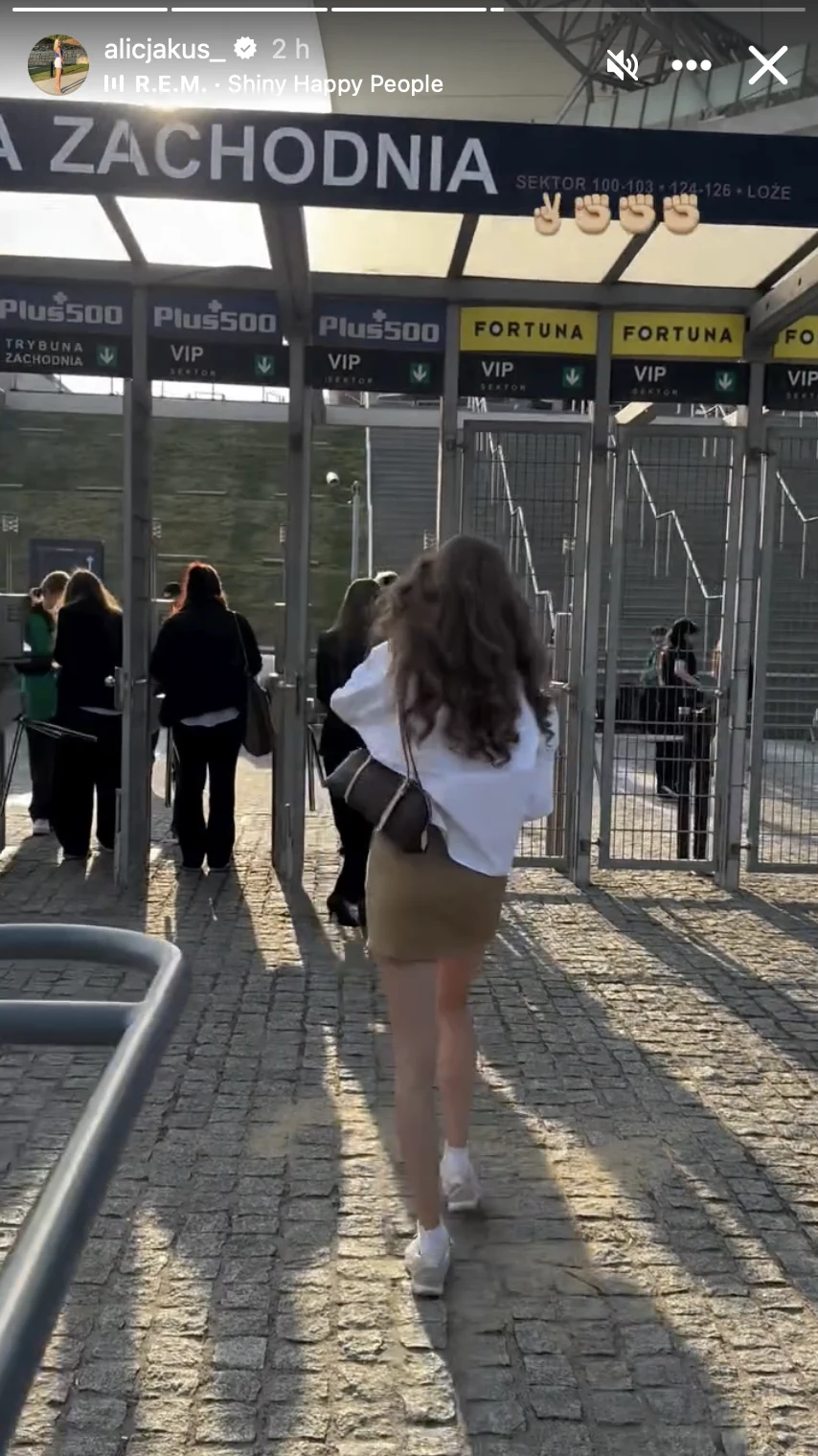 Młoda kobieta w białej bluzie, beżowej spódnicy i białych butach idzie w stronę wejścia VIP na stadion, za którym widać kilka osób oczekujących przy bramkach oraz tablice z nazwami sektorów.
