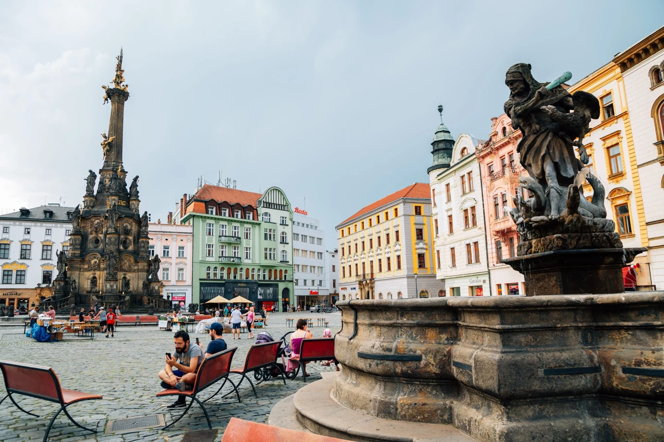 Rynek z kolorowymi kamienicami, monumentalną kolumną i barokową fontanną, wokół której odpoczywają ludzie na ławkach.
