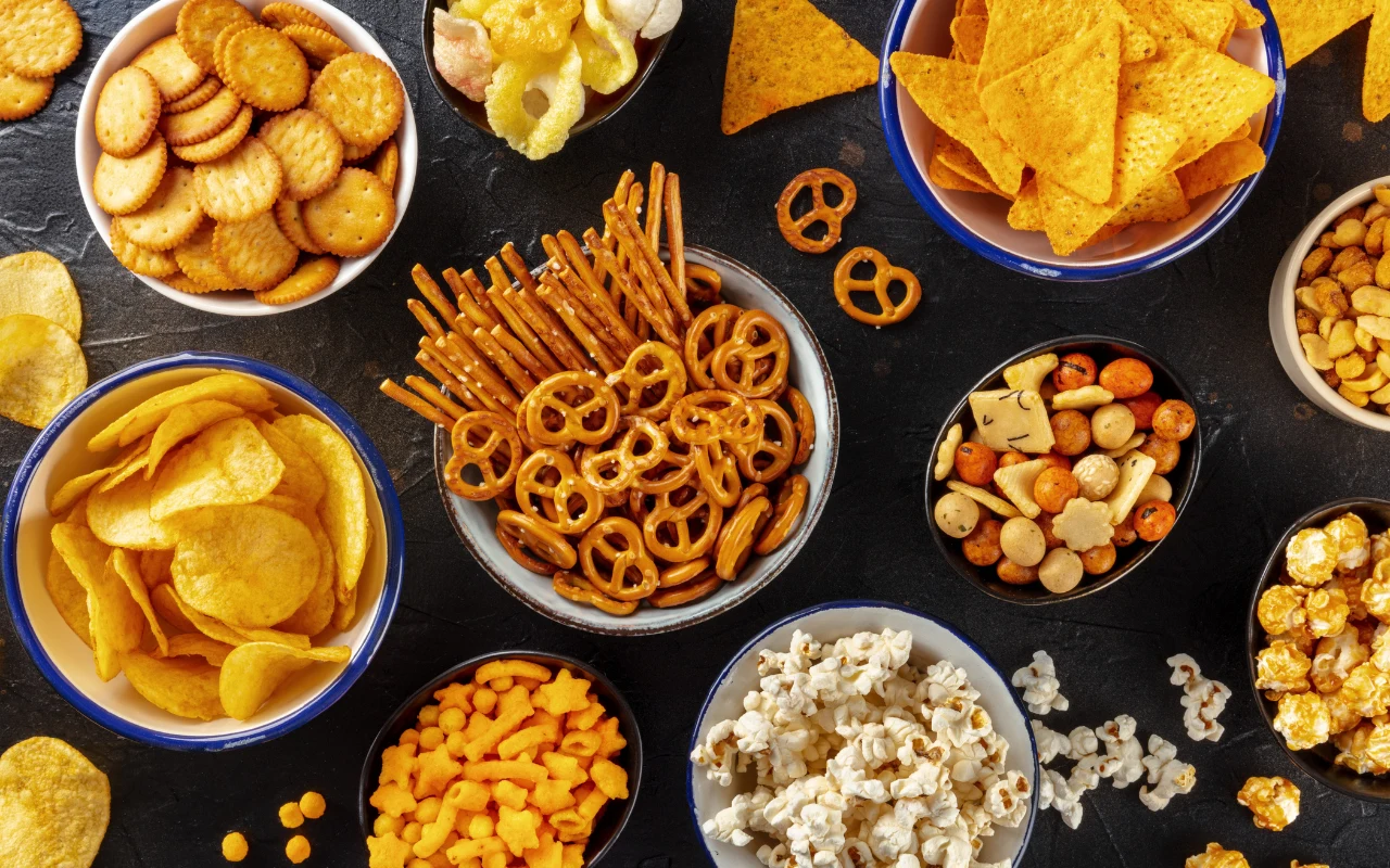 Różnorodne przekąski w miseczkach, w tym krakersy, chipsy ziemniaczane, paluszki, nachosy, popcorn, słone orzeszki i precle, ułożone na ciemnym blacie.