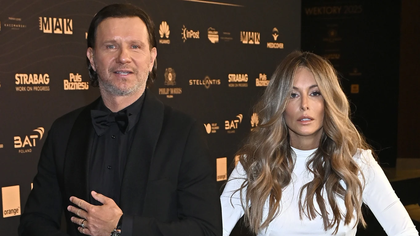 Radosław Majdan, Małgorzata Rozenek-Majdan Elegancko ubrany mężczyzna w czarnym garniturze i kobieta z długimi, falowanymi blond włosami w białej sukni stoją razem na tle ścianki z logotypami partnerów wydarzenia.