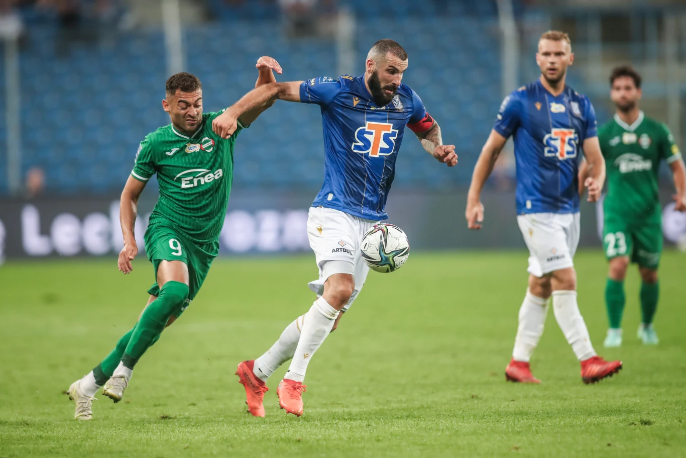 Leandro (z lewej) walczy o piłkę z napastnikiem Lecha Poznań Mikaelem Ishakiem Leandro w zielonym stroju walczy o piłkę z Mikaelem Ishakiem z Lecha Poznań podczas meczu, w tle dwaj gracze.