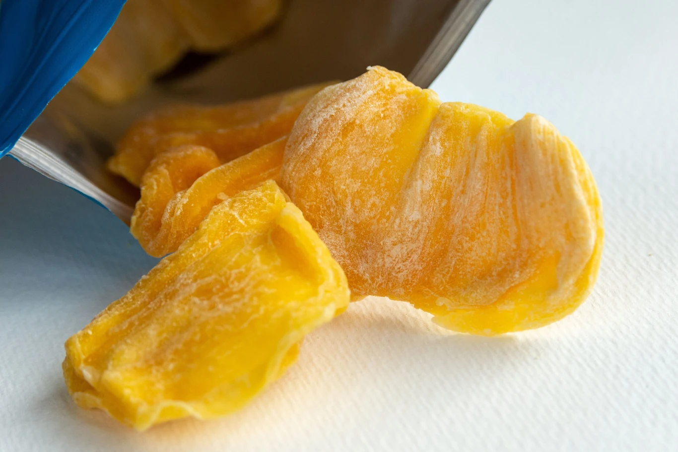 Kilka kawałków suszonego mango wysypanych z opakowania na jasną powierzchnię.