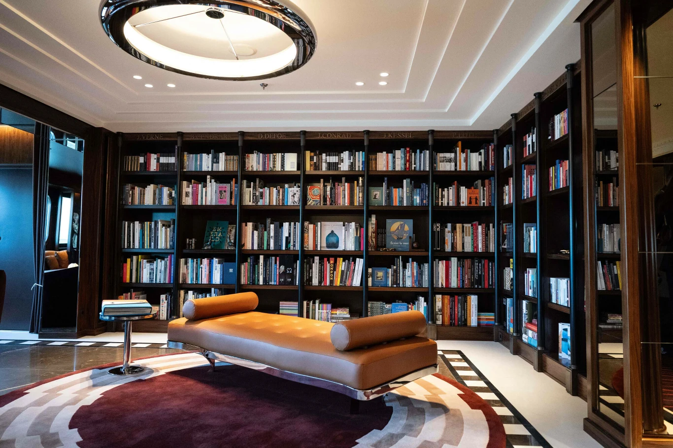 Nowoczesna biblioteka z wysokimi regałami pełnymi książek, centralnie ustawioną skórzaną leżanką oraz designerską lampą sufitową.