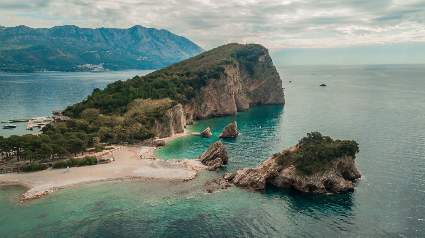 Malownicza zatoka z piaszczystą plażą, otoczona turkusową wodą i skalistymi formacjami porośniętymi zielenią, w tle majestatyczne górskie pasmo pod zachmurzonym niebem.