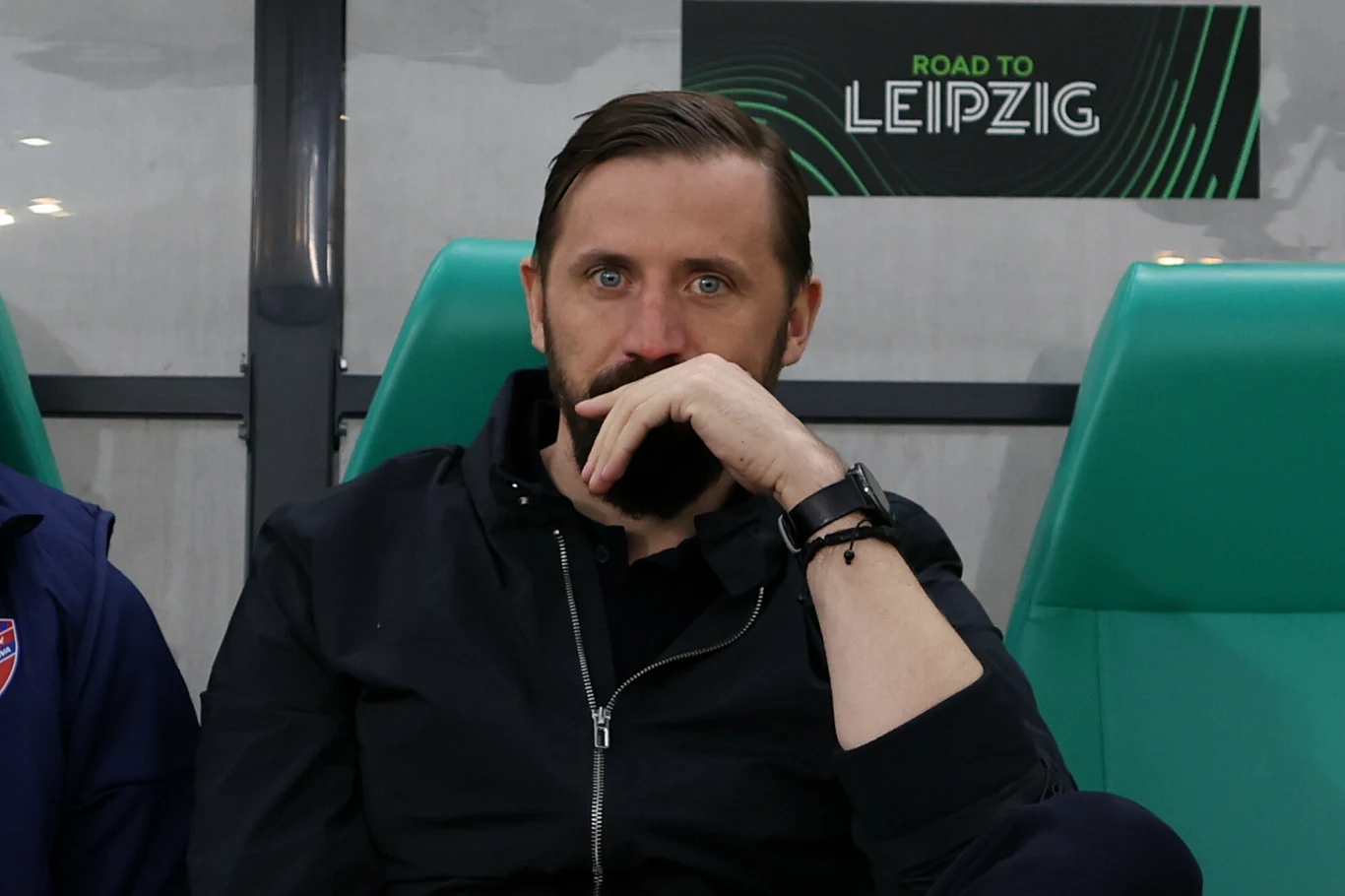 Łukasz Tomczyk Łukasz Tomczyk z brodą i ciemnymi włosami siedzi na zielonym fotelu stadionu, w tle napis 'ROAD TO LEIPZIG'.
