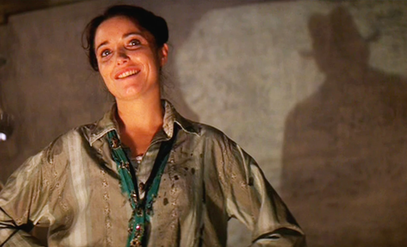 Karen Allen w filmie "Indiana Jones i Poszukiwacze Zaginionej Arki" Karen Allen jako Marion Ravenwood w luźnej koszuli i zielonym naszyjnikiem, obok cień osoby w kapeluszu.