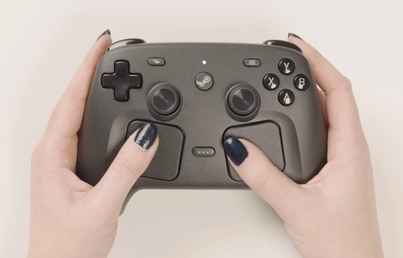 Steam Controller Steam Controller trzymany obiema dłońmi, widoczne przyciski, gałki analogowe i ciemny lakier na paznokciach.