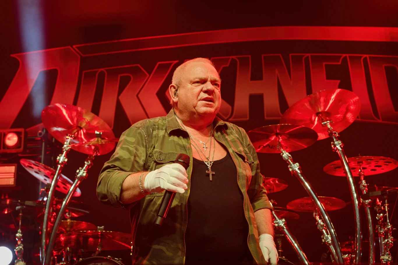 Udo Dirkschneider stoi na czele grupy U.D.O. Udo Dirkschneider w wojskowej koszuli z mikrofonem na scenie przed perkusją i logo zespołu na czerwonym tle.