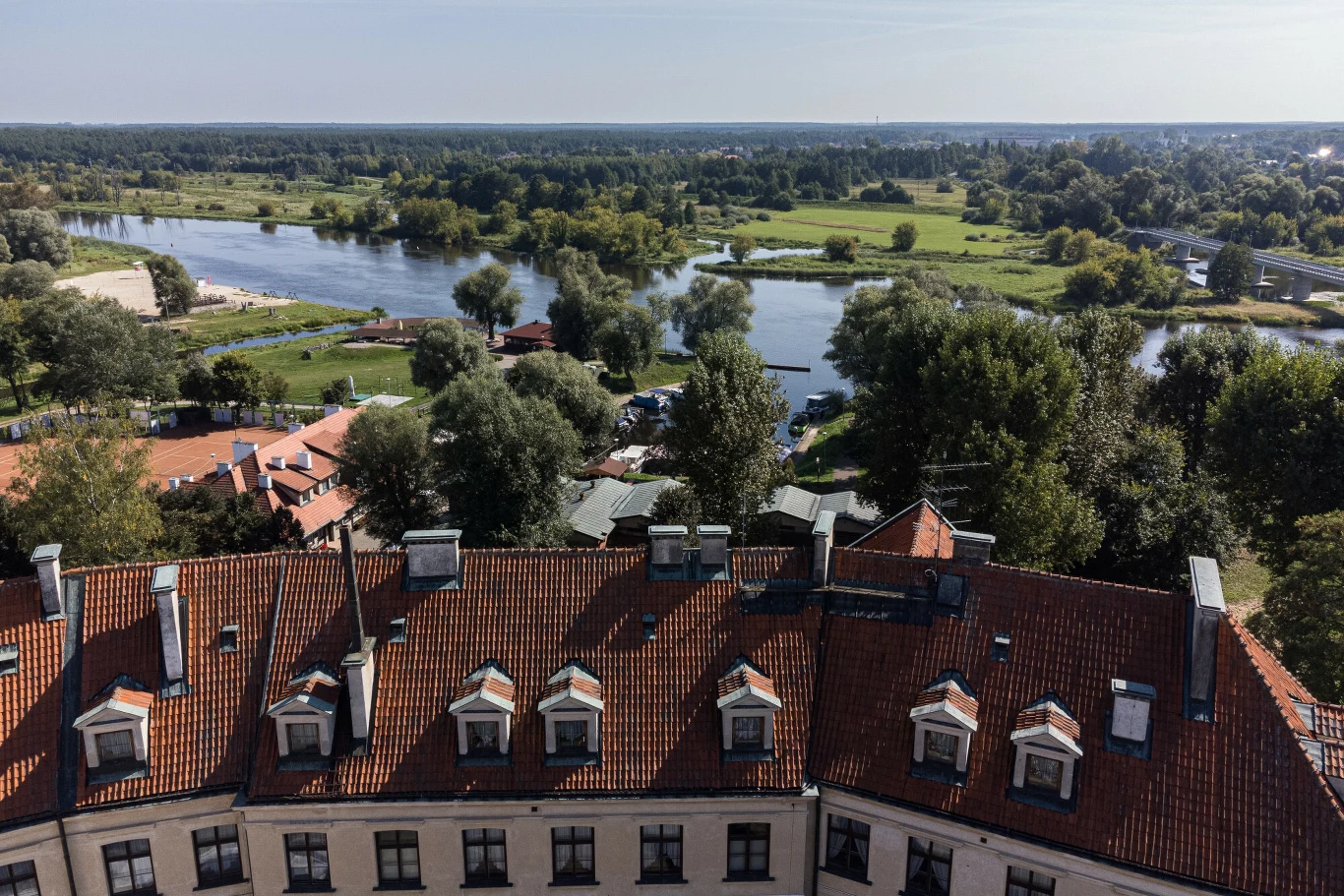 Pułtusk to ”Wenecja Mazowsza” dzięki malowniczym kanałom rzeki Narew Czerwony dach dużego budynku z rzędami mansardowych okien, otoczony zielenią i drzewami, w tle szeroka rzeka wijąca się pośród łąk i lasów.