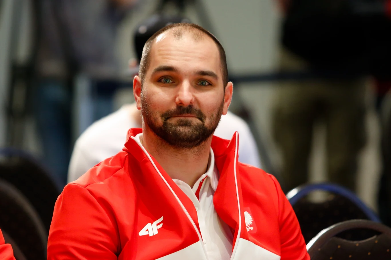Maciej Kurowski Maciej Kurowski w czerwonej sportowej kurtce siedzi na sali wśród innych osób.