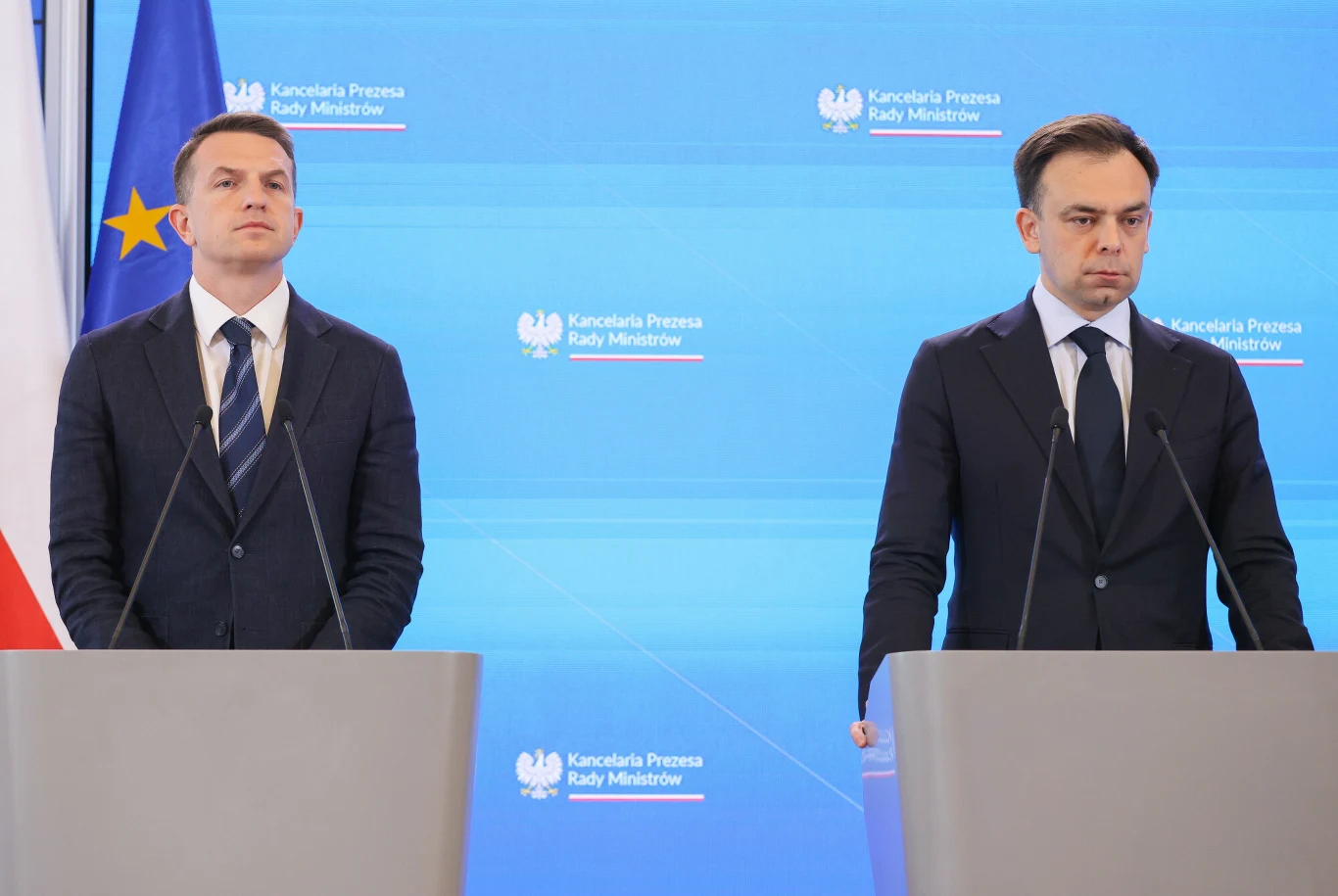Rząd przyjął projekt ustawy o Osobistych Kontach Inwestycyjnych (zdj. ilustracyjne) Minister finansów Andrzej Domański i rzecznik rządu Adam Szłapka w garniturach przy pulpitach na tle logotypów KPRM, widoczne flagi Polski i UE.