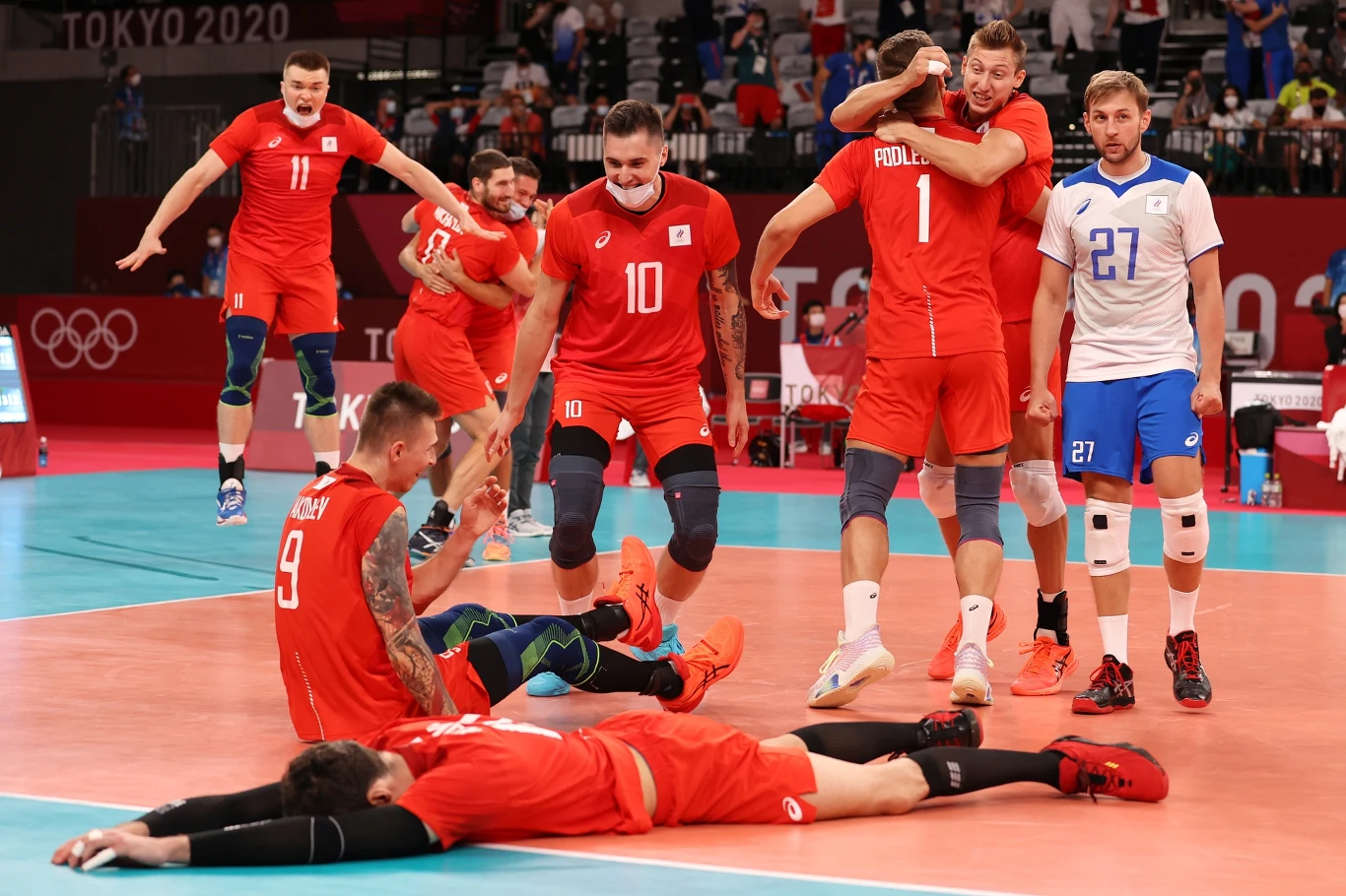 Grupa siatkarzy ubranych na czerwono świętuje zwycięstwo, okazując radość poprzez skakanie, przytulanie i gesty triumfu na boisku do siatkówki, podczas gdy zawodnicy w białych i niebieskich strojach odchodzą w tle.