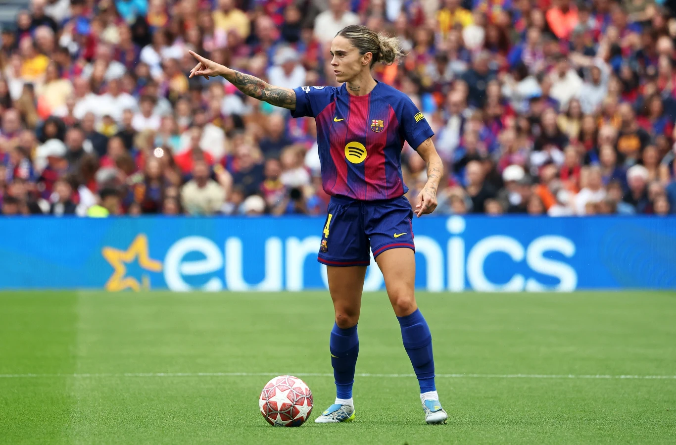 Mapi Leon Piłkarka w stroju FC Barcelony stoi na murawie obok piłki i wskazuje ręką kierunek gry podczas meczu, w tle widoczna pełna kibiców trybuna.