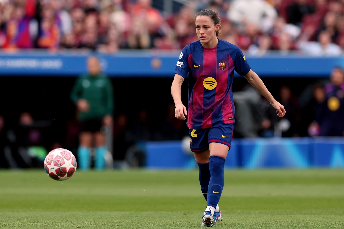 Ona Batlle Piłkarka FC Barcelony w granatowo-bordowym stroju biegnie po murawie z piłką podczas meczu piłkarskiego na stadionie, w tle widoczni kibice na trybunach.