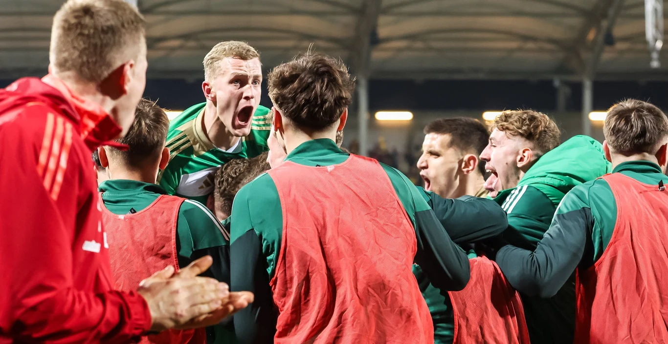Piłkarze ubrani w zielone i czerwone stroje świętują wspólnie zdobycie gola, okazując silne emocje i gesty radości na tle stadionu.