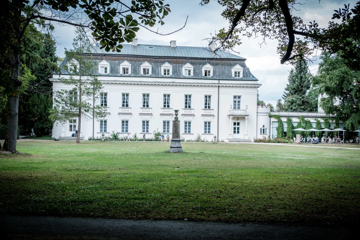 Biała, klasycystyczna rezydencja z wysokimi oknami i mansardowym dachem stoi na środku rozległego, zielonego trawnika, na którym znajduje się pojedynczy pomnik; budynek otaczają wysokie drzewa i subtelne ozdoby pnączy przy bocznym tarasie.