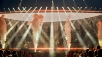 Zespół muzyczny występuje na dużej scenie z efektami świetlnymi i strumieniami dymu, a tłum ludzi ogląda koncert z bliska.