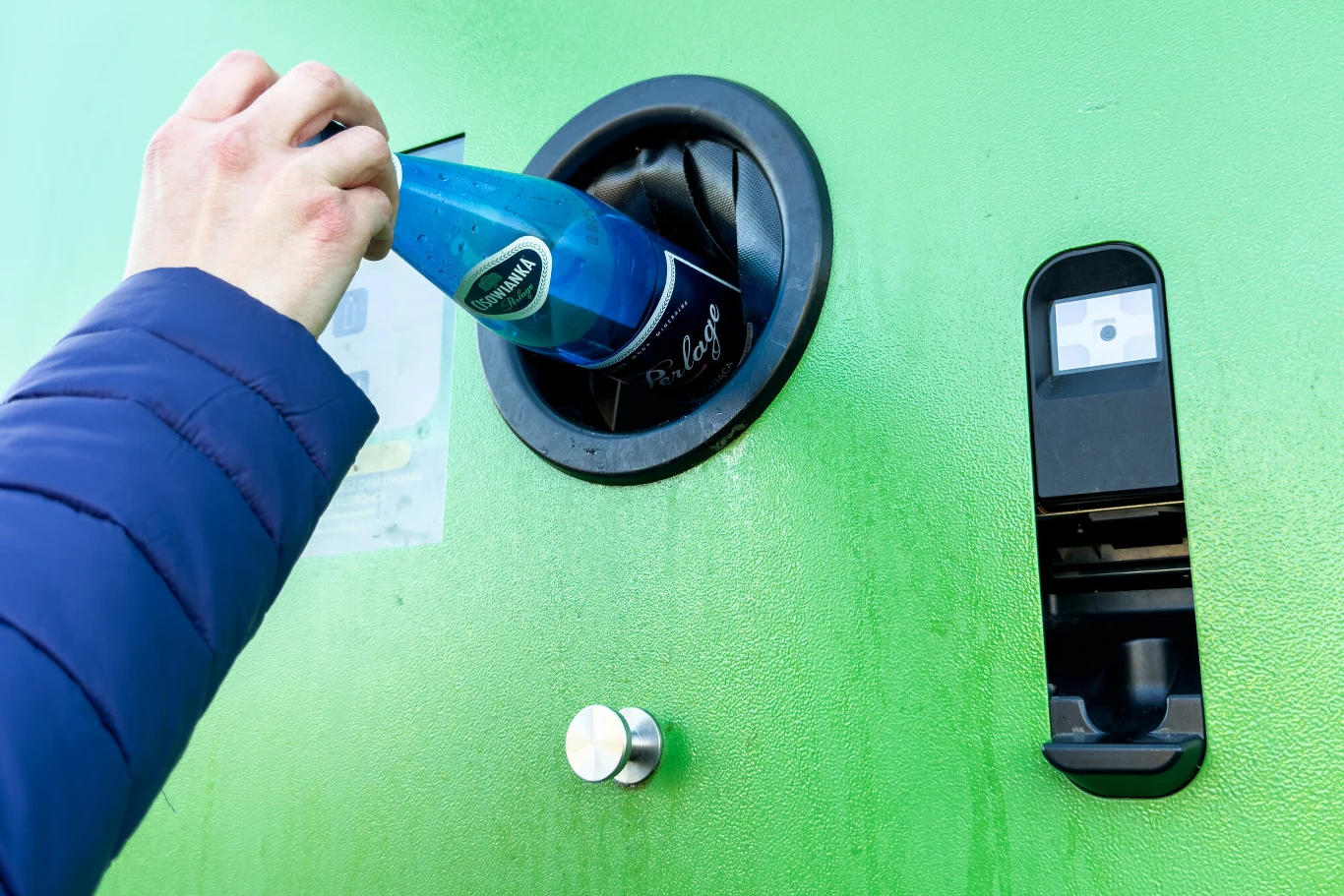 Badania wskazują, że aż 99,6 proc. Polek i Polaków wie o systemie kaucyjnym Osoba w niebieskiej kurtce wkłada plastikową butelkę do automatu do recyklingu umieszczonego na zielonej ścianie.